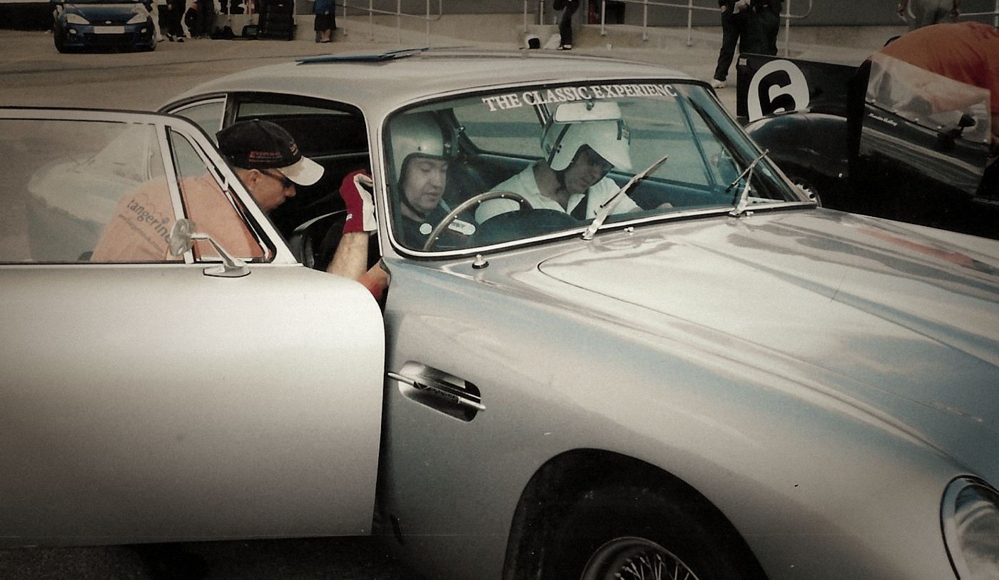Two helmeted drivers in a silver classic Aston Martin in the paddock