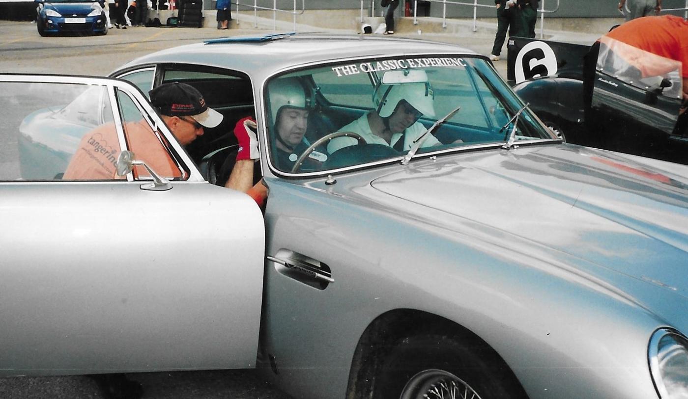 Two helmeted drivers in a silver classic Aston Martin in the paddock